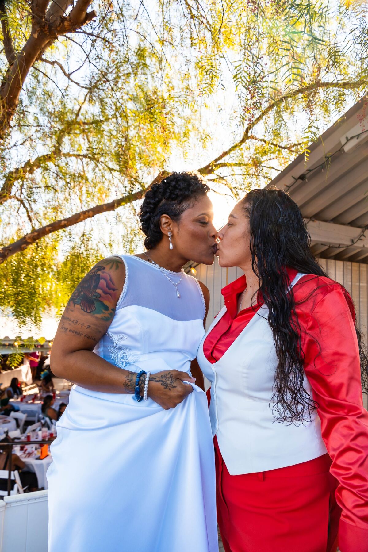 Newlyweds kissing beneath sunlit trees at their red-and-white wedding